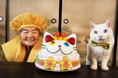 伊原美代子《みさおとふくまる》2012 ⒸMiyoko Ihara　中心にはふくまるの顔の形をしたケーキ。伊原さんが手作りしたもので、みさおさんの米寿をお祝いするもの。「88」の形をしたキャンドル、一字ずつ星の中に書かれた「米」「寿」の文字。ふくまると同じ、右目がイエロー・左目がブルーのオッドアイ。向かって左側には頭巾とちゃんちゃんこを着て笑顔のみさおさん。右側には座卓に両前足をついて身を乗り出すふくまる。首元にはゴールドの蝶ネクタイ